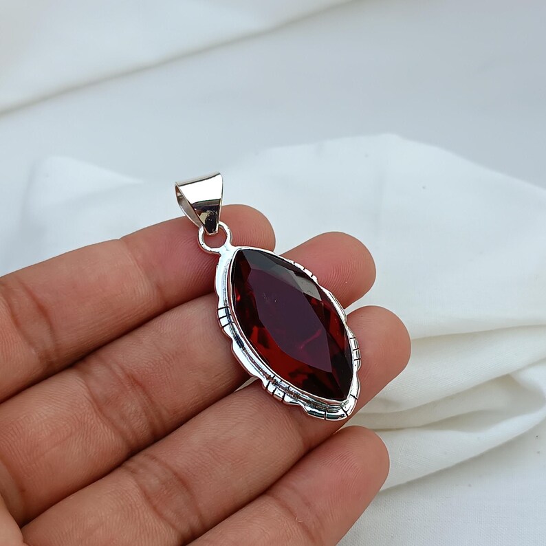 May include: A silver pendant with a red gemstone in a marquise shape. The pendant has a detailed silver border.