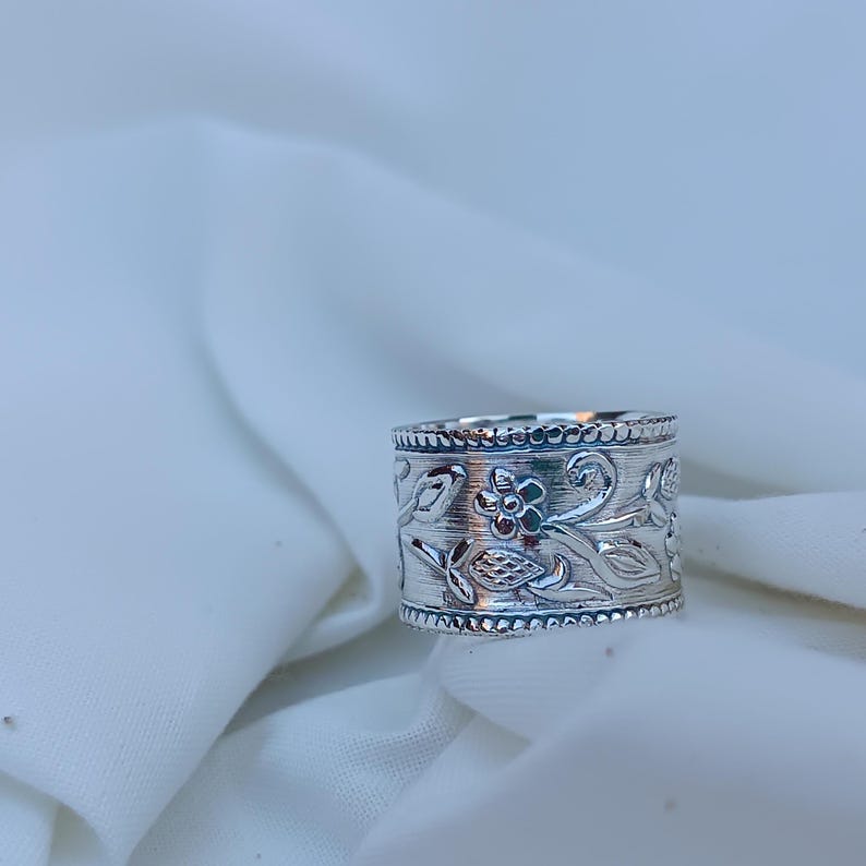 May include: A silver ring with an intricate floral design. The ring is wide and has a textured surface.