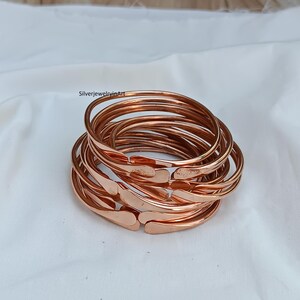 May include: A stack of 10 copper bangles, each with a slightly rounded edge. The bangles are arranged in a circular pattern, with the top bangle slightly offset from the others. The bangles are a warm copper color.