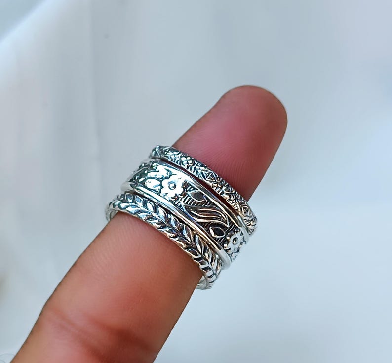 Peut inclure: Une bague en argent orn&eacute;e de motifs floraux et de feuilles complexes. La bague est compos&eacute;e de plusieurs anneaux, chacun avec un design unique. La bague est port&eacute;e sur un doigt, mettant en valeur le savoir-faire d&eacute;taill&eacute; et la surface brillante du m&eacute;tal.