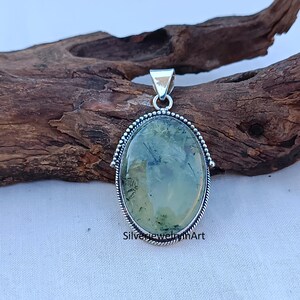 May include: A silver pendant featuring a green and black moss agate stone set in a silver oval frame. The pendant is on a wooden surface.