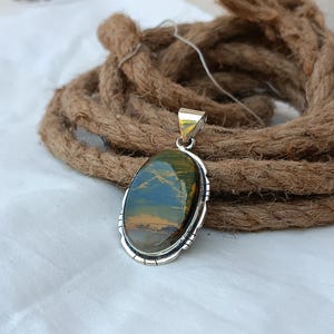 May include: A silver pendant with an oval-shaped tiger's eye gemstone. The gemstone has a green, blue, and gold color pattern. The pendant has a decorative silver border.