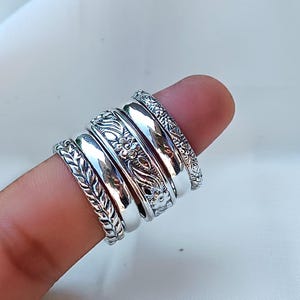 May include: A collection of silver rings, each with a unique design, including floral and leaf patterns. The rings are stacked on a finger, showcasing their various textures and styles. The rings appear to be made of a shiny metal.