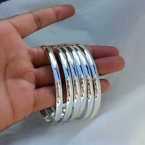 May include: A set of five silver bangles, stacked and displayed on a hand. The bangles are smooth and shiny, with a rounded profile.