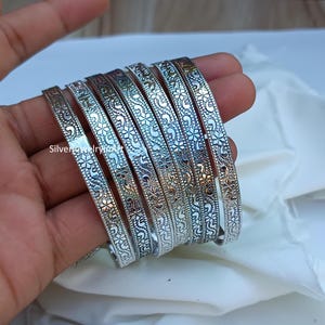 May include: A set of eight silver bangles with a floral and swirl design. The bangles are stacked on top of each other.