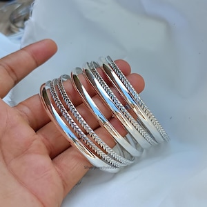 May include: A set of six silver bangles with a braided design. The bangles are stacked on top of each other and are all the same size.