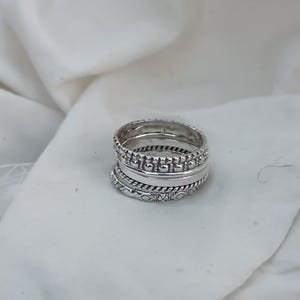 Könnte beinhalten: Ein Stapel silberner Ringe mit verschiedenen Texturen. Die Ringe weisen Perlen-, glatte und gemusterte Designs auf. Die Ringe werden auf einer weißen, strukturierten Oberfläche präsentiert.