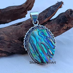 Könnte beinhalten: Ein silberner Anhänger mit einem ovalen Einlegeteil aus Abalone-Muschel. Die Muschel hat ein blaues, grünes und violettes irisierendes Muster. Der Anhänger ist von einem silbernen Rand mit einem dekorativen Design umgeben.