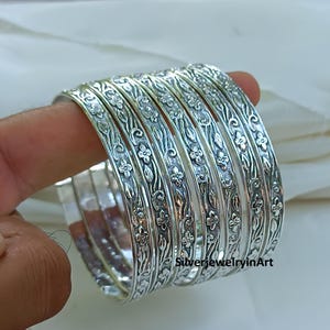 Op de afbeelding: Een stapel zilveren armbanden met bloemen- en bladerpatronen. De armbanden zijn op elkaar gestapeld en het bloemmotief is zichtbaar op het oppervlak. De sieraden worden op een witte achtergrond gepresenteerd.