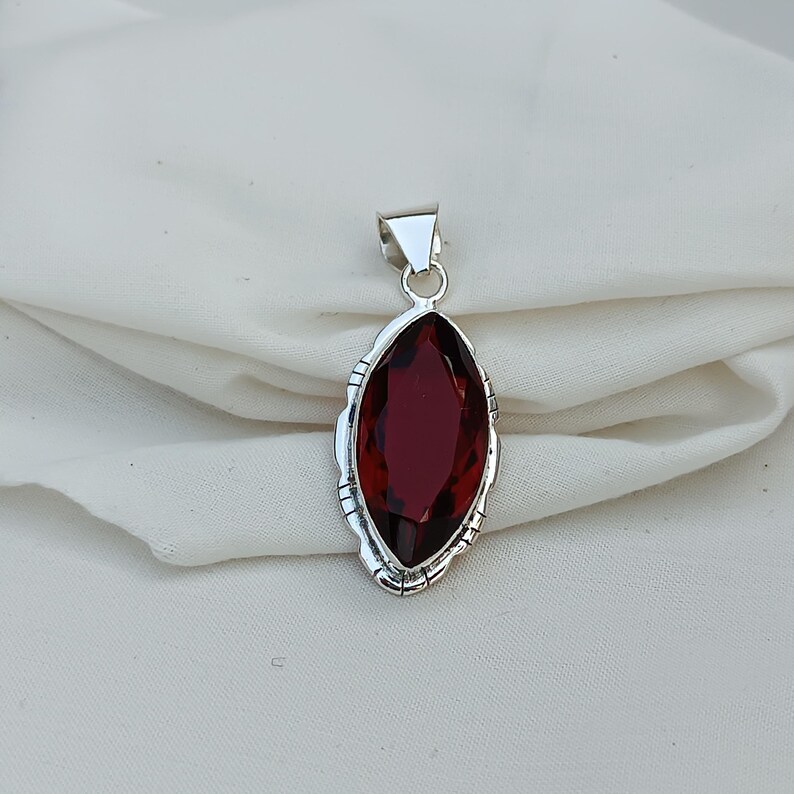 May include: A silver pendant with a dark red gemstone in a marquise shape. The pendant has a detailed, ornate design around the gemstone.