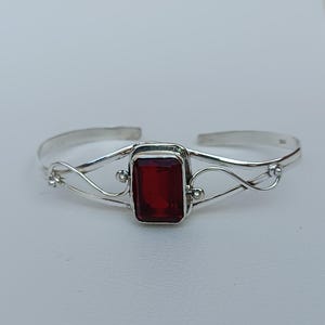 May include: A silver bangle bracelet with a red gemstone set in a square frame. The bracelet has a delicate, intricate design.