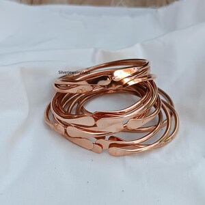 May include: A stack of 10 copper bangles, each with a slightly different width and a rounded edge. The bangles are arranged in a loose pile, with the top bangle slightly offset from the others.