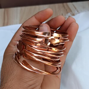 May include: A stack of 10 copper bangles, each with a slightly different shape and width. The bangles are smooth and shiny, and they are stacked on top of each other in a hand.