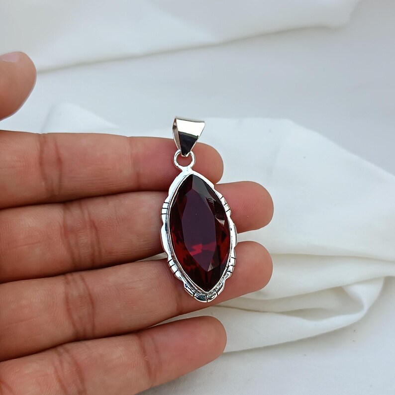 May include: A silver pendant with a red gemstone in a marquise shape. The pendant has a detailed silver border.