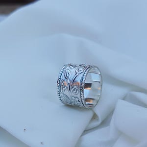 May include: A silver ring with a floral design. The ring is wide and has a textured surface.