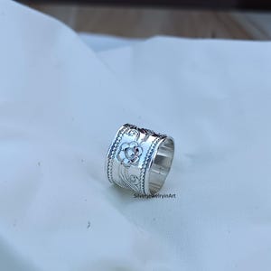 Op de afbeelding: Een zilveren ring met een bloemmotief. De ring is breed en heeft een getextureerd oppervlak.