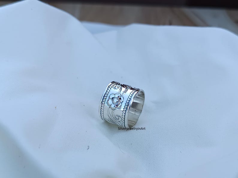 Op de afbeelding: Een zilveren ring met een bloemmotief. De ring is breed en heeft een getextureerd oppervlak.
