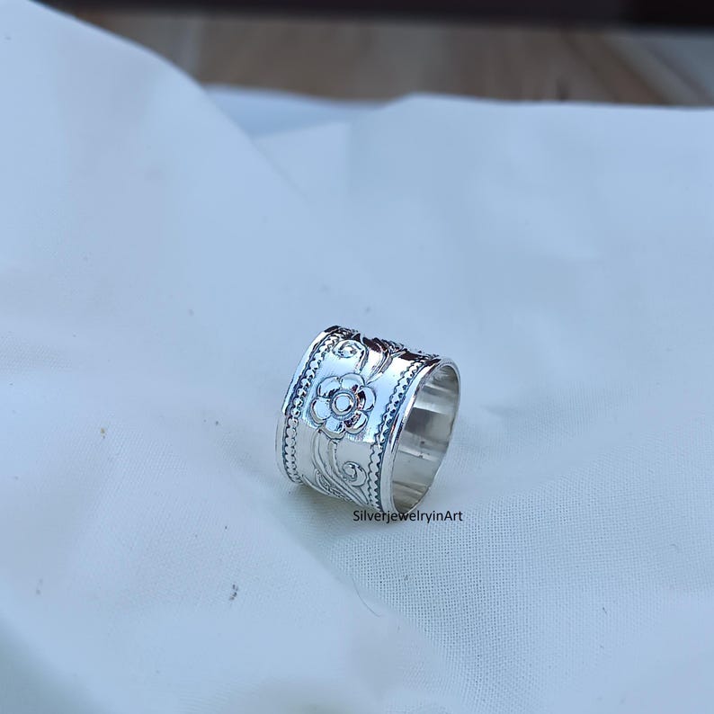 Op de afbeelding: Een zilveren ring met een ingewikkeld bloemmotief. De ring is breed en heeft een getextureerd oppervlak.