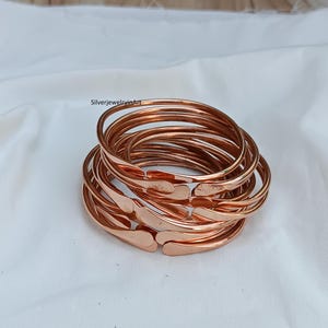 May include: A stack of ten copper bangles, each with a slightly hammered texture. The bangles are arranged in a loose pile, with the ends of the bangles overlapping.