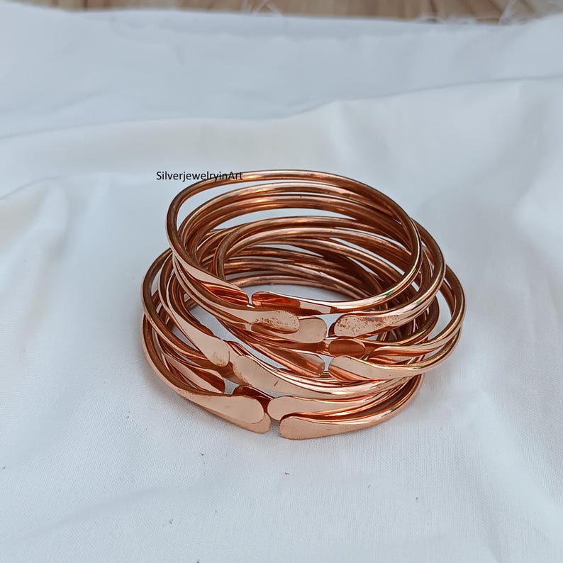May include: A stack of ten copper bangles, each with a slightly hammered texture. The bangles are arranged in a loose pile, with the ends of the bangles overlapping.