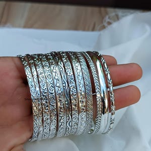 Può includere: Un set di braccialetti d'argento con vari disegni in rilievo. I braccialetti sono impilati uno sopra l'altro e hanno una finitura lucida.