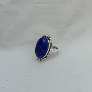 Op de afbeelding: Een zilveren ring met een grote ovale lapis lazuli-edelsteen. De ring heeft een gedetailleerde zilveren band met een parelrand.