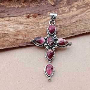 Könnte beinhalten: Silberfarbener Kreuzanhänger mit tropfenförmigen, weinroten Steinen und Goldflocken. Das Kreuz hat eine dekorative Öse und ist vor einem Holzuntergrund platziert. Ein religiöses oder spirituelles Accessoire.