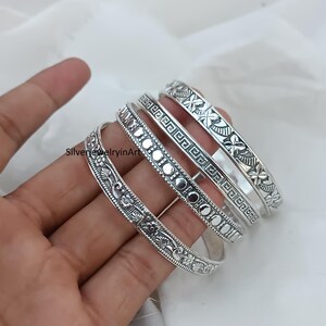 Könnte beinhalten: Set aus vier silbernen Armreifen mit aufwendigen floralen und geometrischen Mustern. Die Armreifen sind übereinander gestapelt und zeigen die verschiedenen Muster.