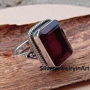 Peut inclure: Une bague en argent avec une grosse pierre précieuse rectangulaire rouge foncé. La pierre est sertie dans une lunette en argent avec un motif en forme de corde sur les bords. La bague est sur une surface en bois. Le texte "SilverjewelryinArt" est visible.