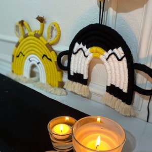 May include: Two macrame rainbows, one yellow and white, the other black and white, with a yellow stripe, hanging on a white wall. Three glass candle holders with lit candles are on a black surface in the foreground.