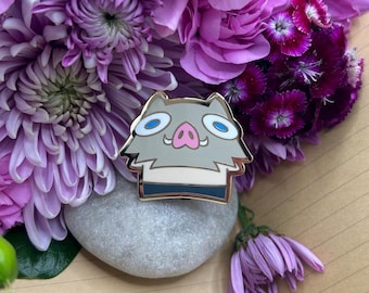 May include: A gold-colored enamel pin featuring a cartoon boar with blue eyes, a pink nose, and a white stripe across its forehead. The boar is wearing a black and white striped shirt.