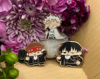 May include: Three enamel pins featuring cartoon characters. The characters are wearing black and white outfits. The pins are gold-coloured and have a shiny finish.