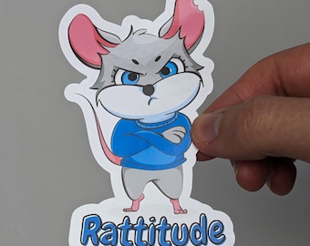 Op de afbeelding: Een gestanste vinylsticker met een grijze cartoonmuis met blauwe ogen, gekleed in een blauwe trui met de armen over elkaar en een boze uitdrukking. De muis heeft roze binnenoren en een lange staart. Onderaan staat het woord Rattitude in het blauw.