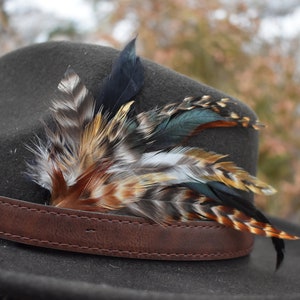 Op de afbeelding: Een grijze wollen fedora hoed met een bruin leren band en een verenversiering. De veren zijn veelkleurig, waaronder zwart, bruin, wit en teal.