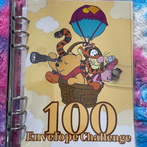 Op de afbeelding: Een doorzichtige binder met een gele kaft met Winnie de Poeh, Teigetje en Knorretje in een mand onder een parachute. De kaft leest "100 Envelope Challenge" en "DONT OVERTHINK ART".