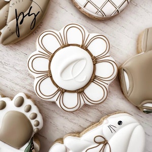 K&ouml;nnte beinhalten: Eine Sammlung von dekorierten Zuckerkeksen mit verschiedenen Designs, darunter eine Blume, ein Hase, ein Pfotenabdruck und ein geometrisches Muster. Die Kekse sind mit wei&szlig;em und braunem Zuckerguss verziert und der Text "@paisleesweets" ist auf einem der Kekse sichtbar.