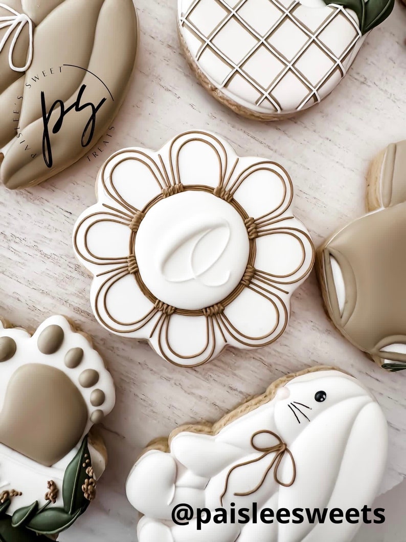 K&ouml;nnte beinhalten: Eine Sammlung von dekorierten Zuckerkeksen mit verschiedenen Designs, darunter eine Blume, ein Hase, ein Pfotenabdruck und ein geometrisches Muster. Die Kekse sind mit wei&szlig;em und braunem Zuckerguss verziert und der Text "@paisleesweets" ist auf einem der Kekse sichtbar.