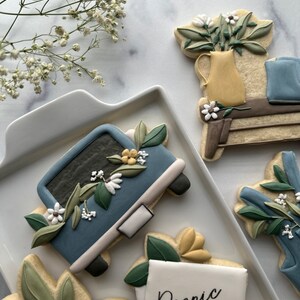 May include: A set of five decorated sugar cookies featuring a blue truck with flowers, a floral arrangement in a yellow vase, a note that says "Picnic at 4 pm", and two additional floral designs.