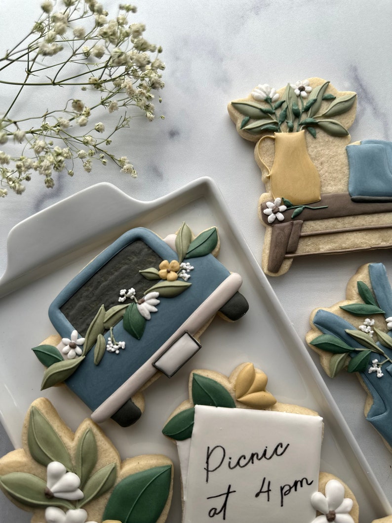 May include: A set of five decorated sugar cookies featuring a blue truck with flowers, a floral arrangement in a yellow vase, a note that says "Picnic at 4 pm", and two additional floral designs.