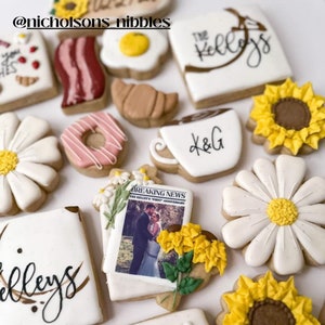 May include: Assortment of decorated sugar cookies with various designs, including a newspaper, a coffee cup, a donut, a daisy, a sunflower, and the text "The Kelleys".