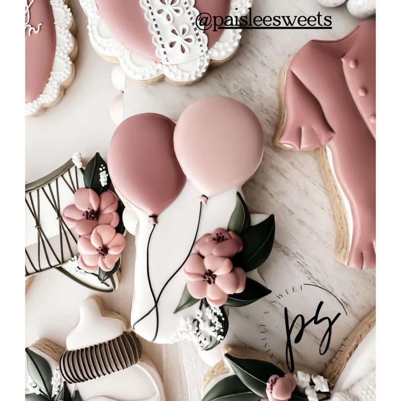 May include: A set of decorated sugar cookies featuring a pair of pink balloons, a floral design, a corset, a baby bottle, and a pink shirt. The cookies are decorated with white icing and have a delicate, feminine aesthetic.