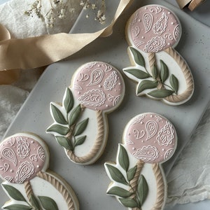 May include: Four decorated sugar cookies shaped like baby rattles with a pink and white paisley design and green leaves. The cookies are on a gray background.