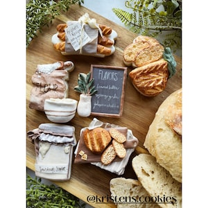 May include: A collection of decorated sugar cookies shaped like bread, flour, and a sourdough starter jar. The cookies are arranged on a wooden surface with a chalkboard sign listing different flavors: browned butter, rosemary garlic, and jalapeno cheddar. The text "Krisher's Recipe" is visible on a piece of parchment paper.