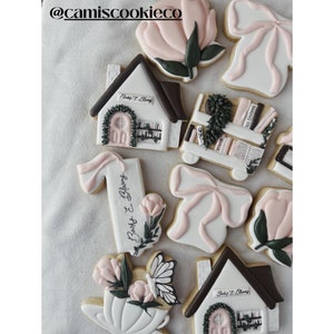May include: A collection of decorated sugar cookies featuring a book and flower theme. The cookies are decorated with pink, white, and green icing and feature images of books, flowers, a teacup, a butterfly, and a house. The cookies are decorated with the text "Books & Blooms".