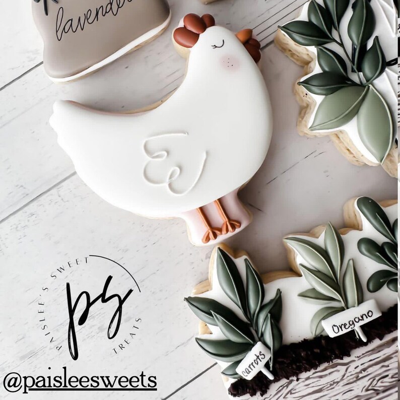 K&ouml;nnte beinhalten: Ein Set dekorierter Zuckerkekse mit einem wei&szlig;en Huhn, einem braunen und wei&szlig;en Keks mit dem Wort "Lavendel" darauf und zwei gr&uuml;nen Keksen mit den Worten "Karotten" und "Oregano" darauf. Die Kekse sind mit wei&szlig;em Zuckerguss verziert und haben einen rustikalen, l&auml;ndlichen Stil.