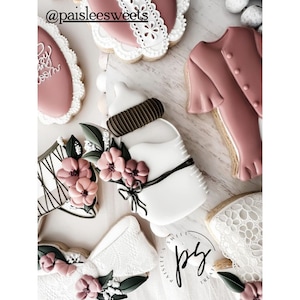 May include: A set of decorated sugar cookies featuring a baby bottle, a bow, a dress, and a corset. The cookies are decorated with white, brown, and pink icing and have a floral theme.