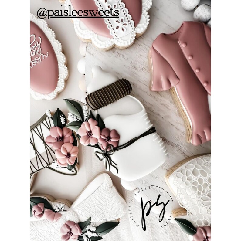 May include: A set of decorated sugar cookies featuring a baby bottle, a bow, a dress, and a corset. The cookies are decorated with white, brown, and pink icing and have a floral theme.