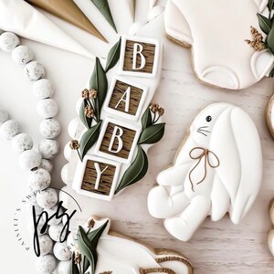 K&ouml;nnte beinhalten: Ein Set dekorierter Zuckerkekse f&uuml;r eine Babyparty. Die Kekse sind in Form eines Stramplers, eines Hasen, eines Korbs und eines Satzes Bl&ouml;cke geformt, die "BABY" buchstabieren. Die Kekse sind mit wei&szlig;em Zuckerguss und gr&uuml;nen Bl&auml;ttern verziert.