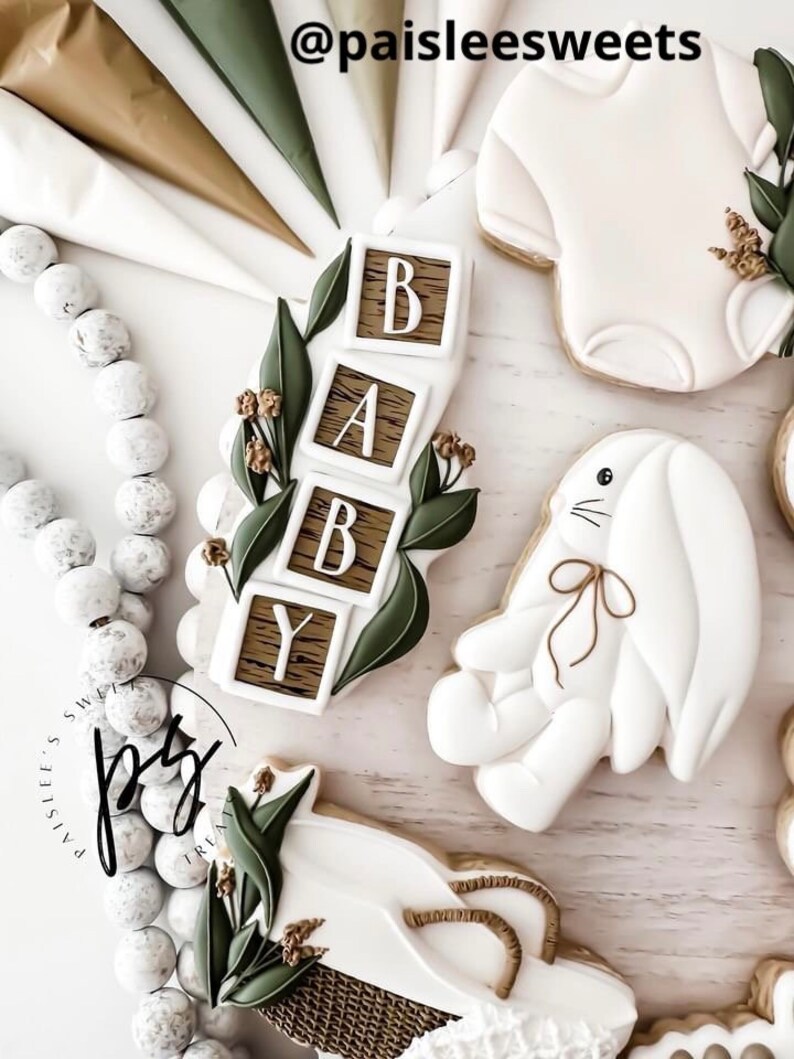 K&ouml;nnte beinhalten: Ein Set dekorierter Zuckerkekse f&uuml;r eine Babyparty. Die Kekse sind in Form eines Stramplers, eines Hasen, eines Korbs und eines Satzes Bl&ouml;cke geformt, die "BABY" buchstabieren. Die Kekse sind mit wei&szlig;em Zuckerguss und gr&uuml;nen Bl&auml;ttern verziert.