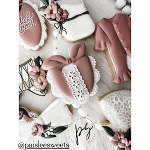 May include: A collection of decorated sugar cookies with various designs, including a baby onesie, a bow, a flower, and a lace pattern. The cookies are decorated in shades of pink, white, and green. The cookies are arranged on a white surface. The text "Baby Bloom" is visible on one of the cookies.